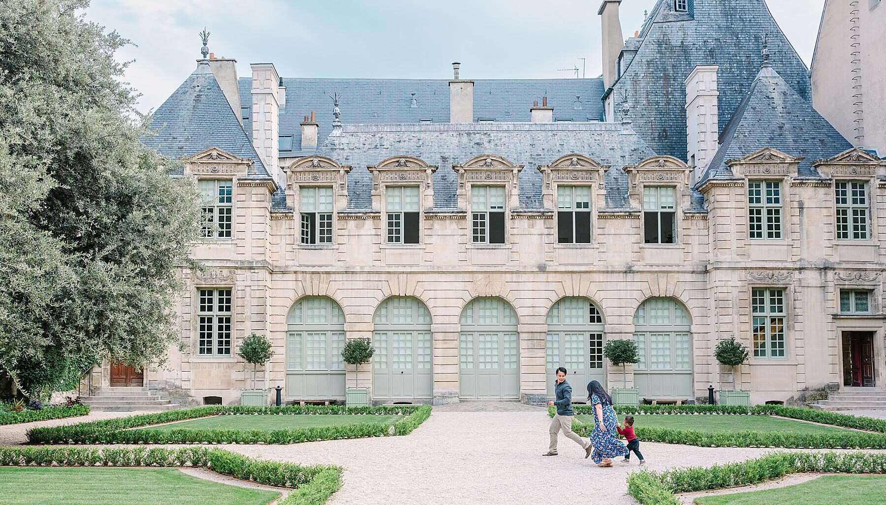 Parents and toddler playing chase through the Hôtel de Sully garden during a family photoshoot in the Marais, Paris