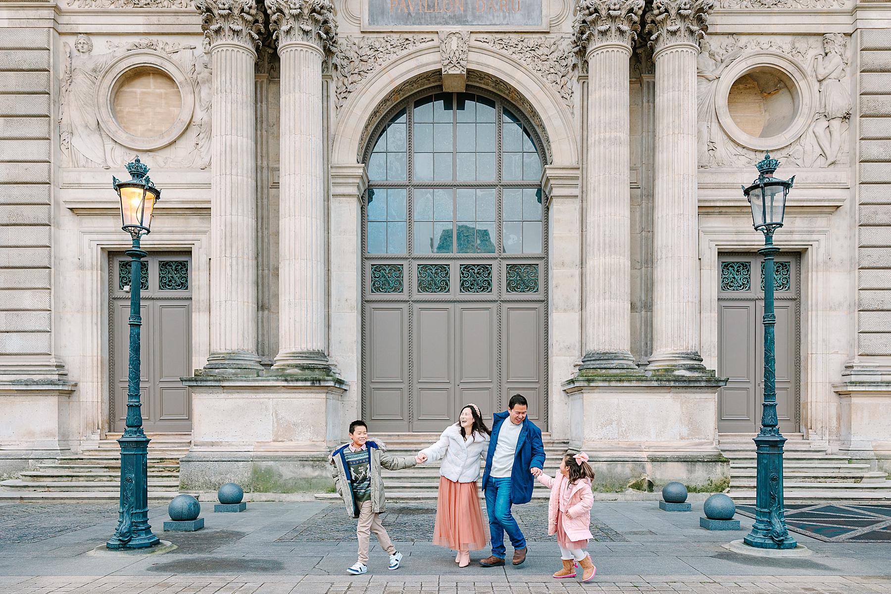 A family of four dressed in coordinating coats and cozy layers walks hand-in-hand in front of the Louvre’s grand architecture during a Paris photoshoot.