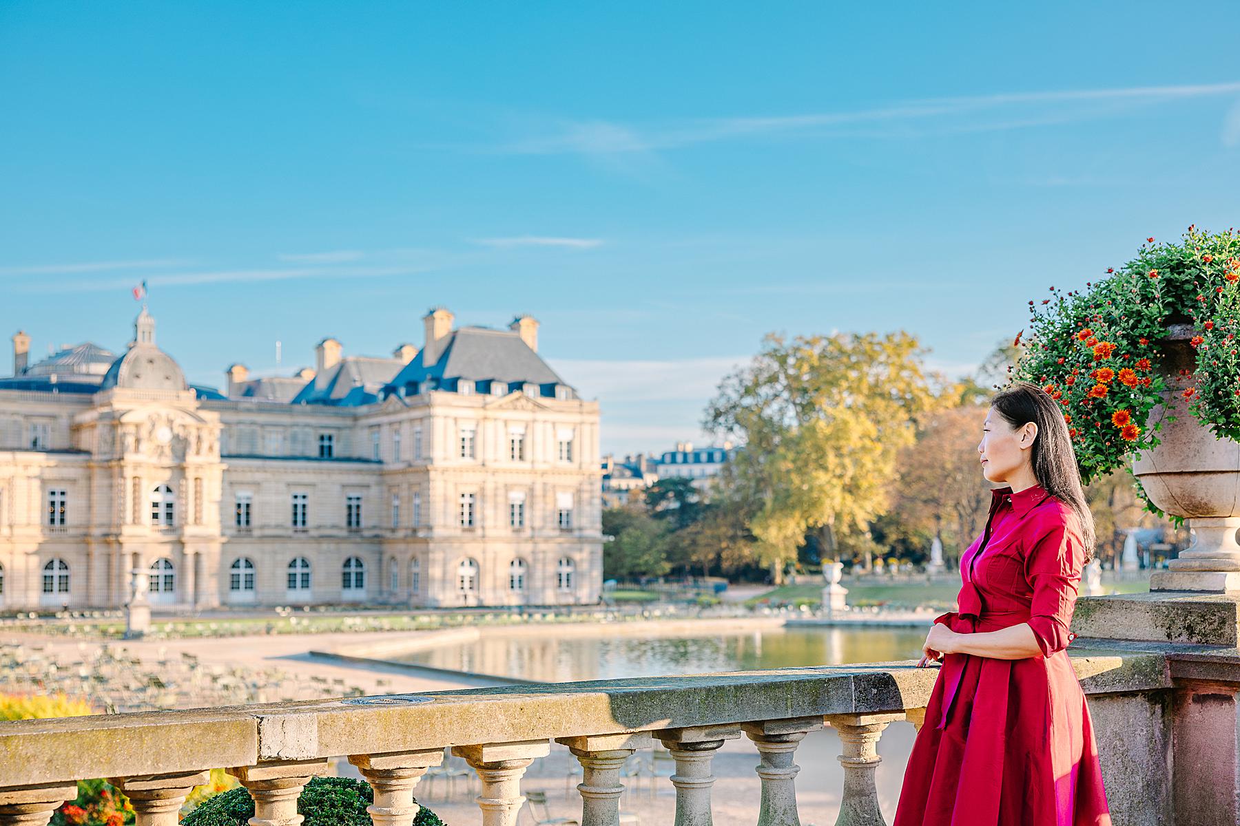 Woman in a red dress standing at a stone railing overlooking the Luxembourg Gardens during a solo photoshoot in the Latin Quarter of Paris