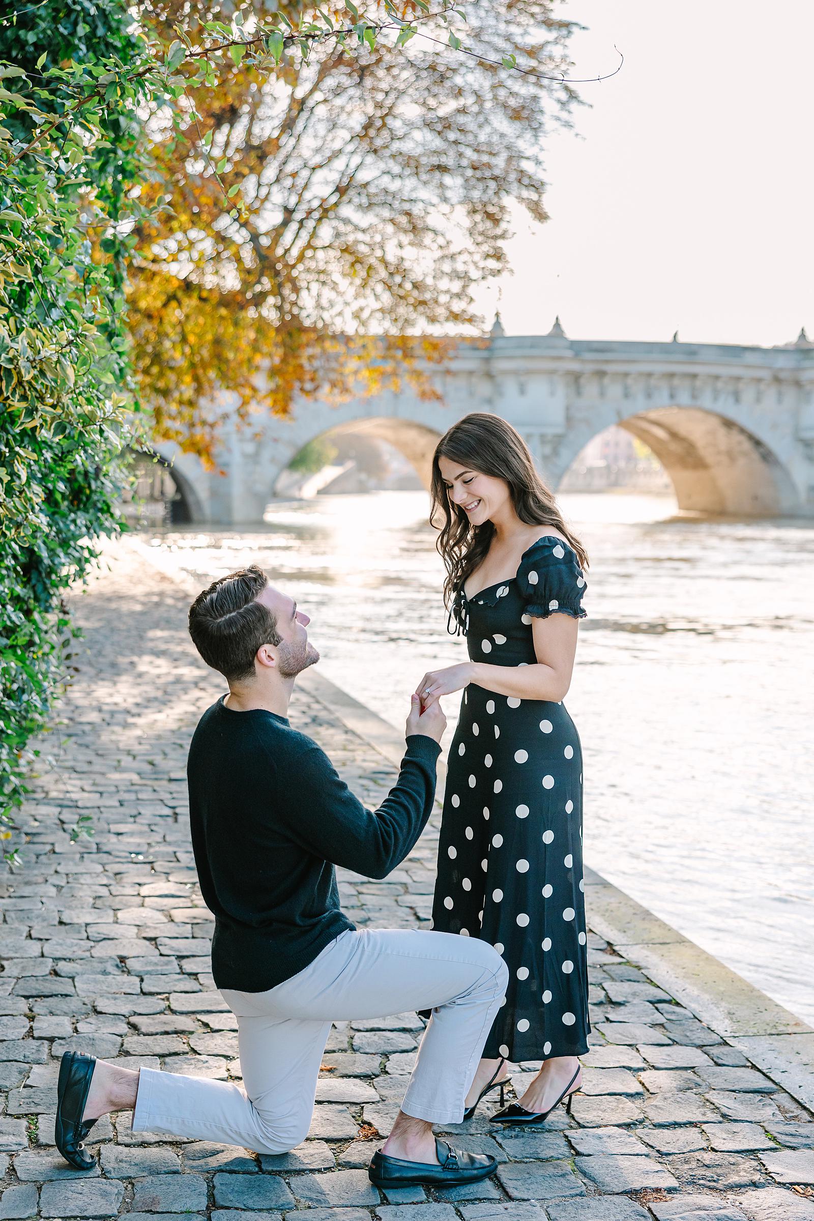 A man on one knee holding a woman's hands, both smiling joyfully after a proposal on the Île de la Cité riverbank, with a bridge in the background.