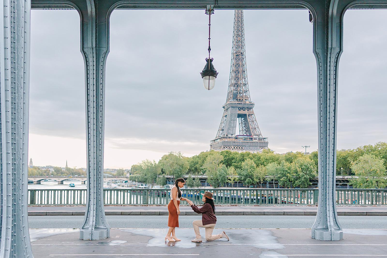 One woman on one knee proposing to another woman on the Bir-Hakeim Bridge, framed by its arches, with the Eiffel Tower prominently in the background.