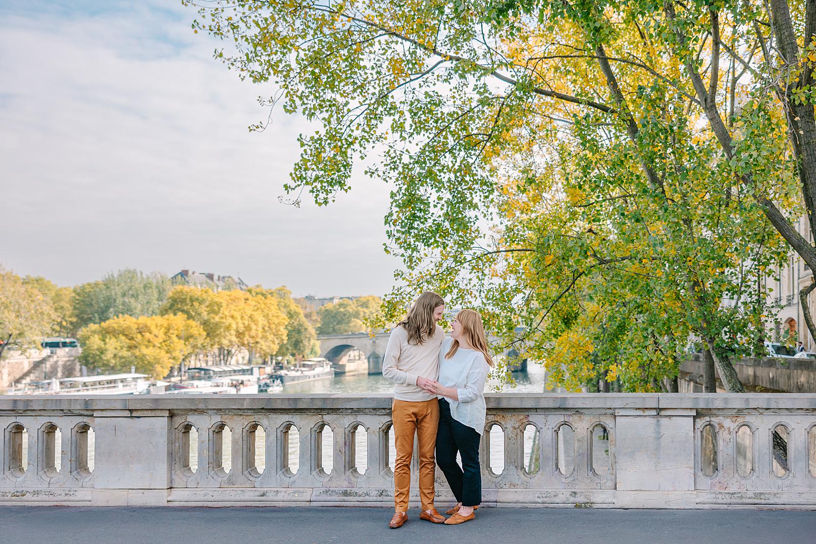 A couple standing close together on a bridge railing on Île Saint-Louis, looking at each other, with the Seine River and autumnal trees and bridges in the background.