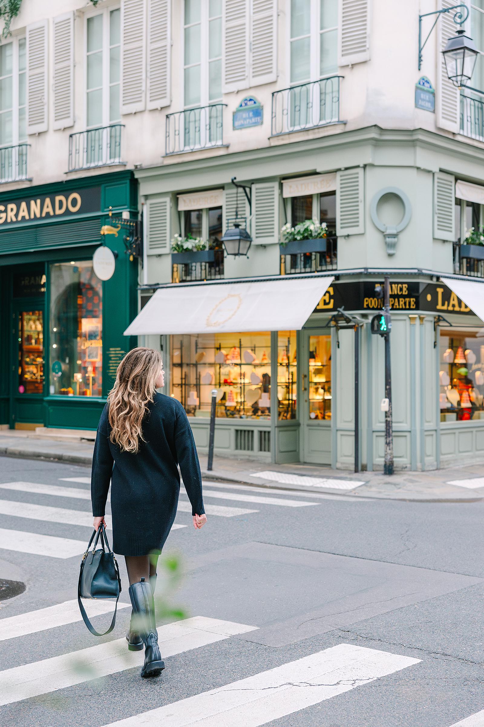 A woman in a dark coat and boots walking across a crosswalk on a street corner in the Latin Quarter, with traditional Parisian buildings and shops behind her.