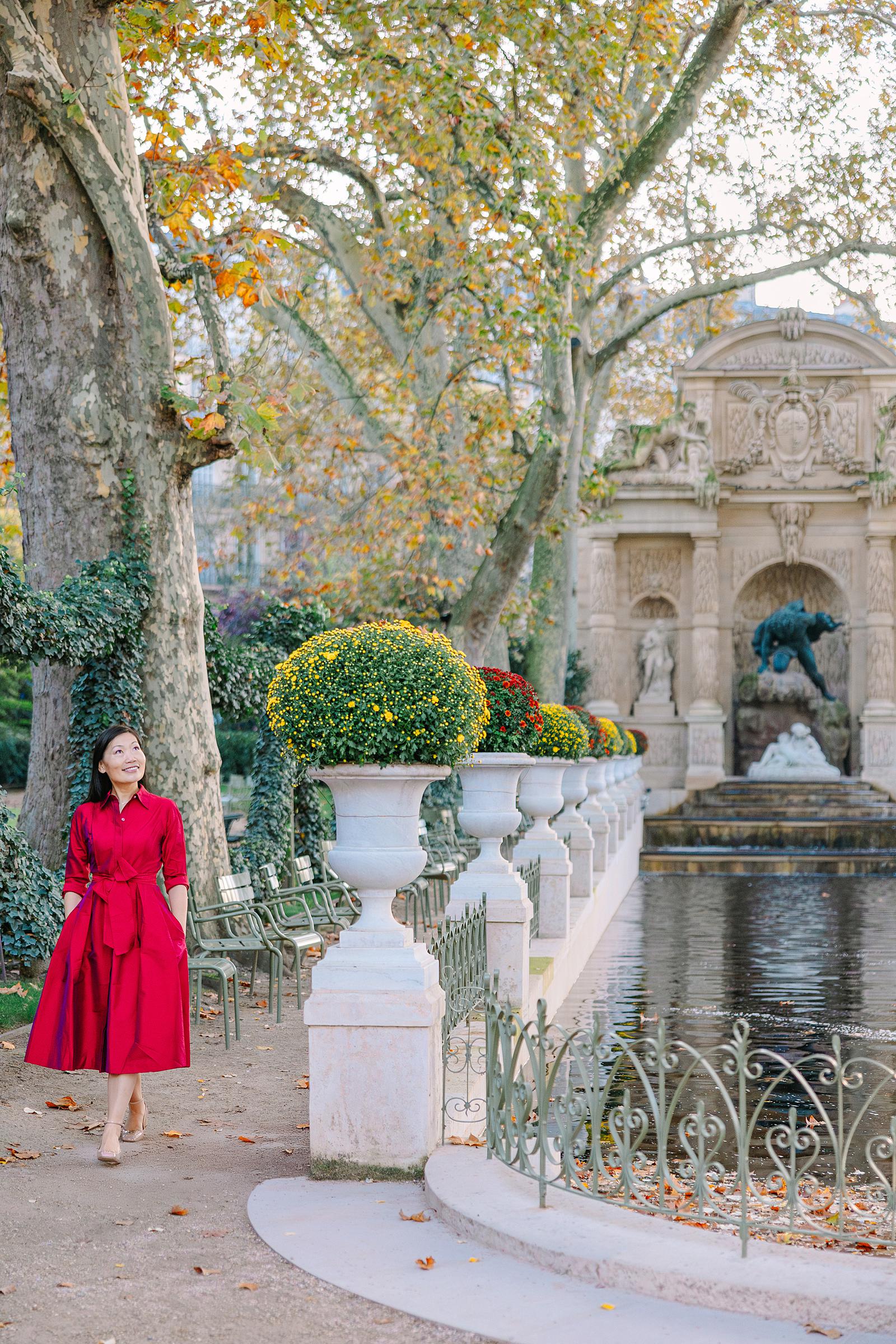 A smiling woman in a red dress walking along a path next to the Medici Fountain in the Luxembourg Gardens, surrounded by autumn trees and planters.