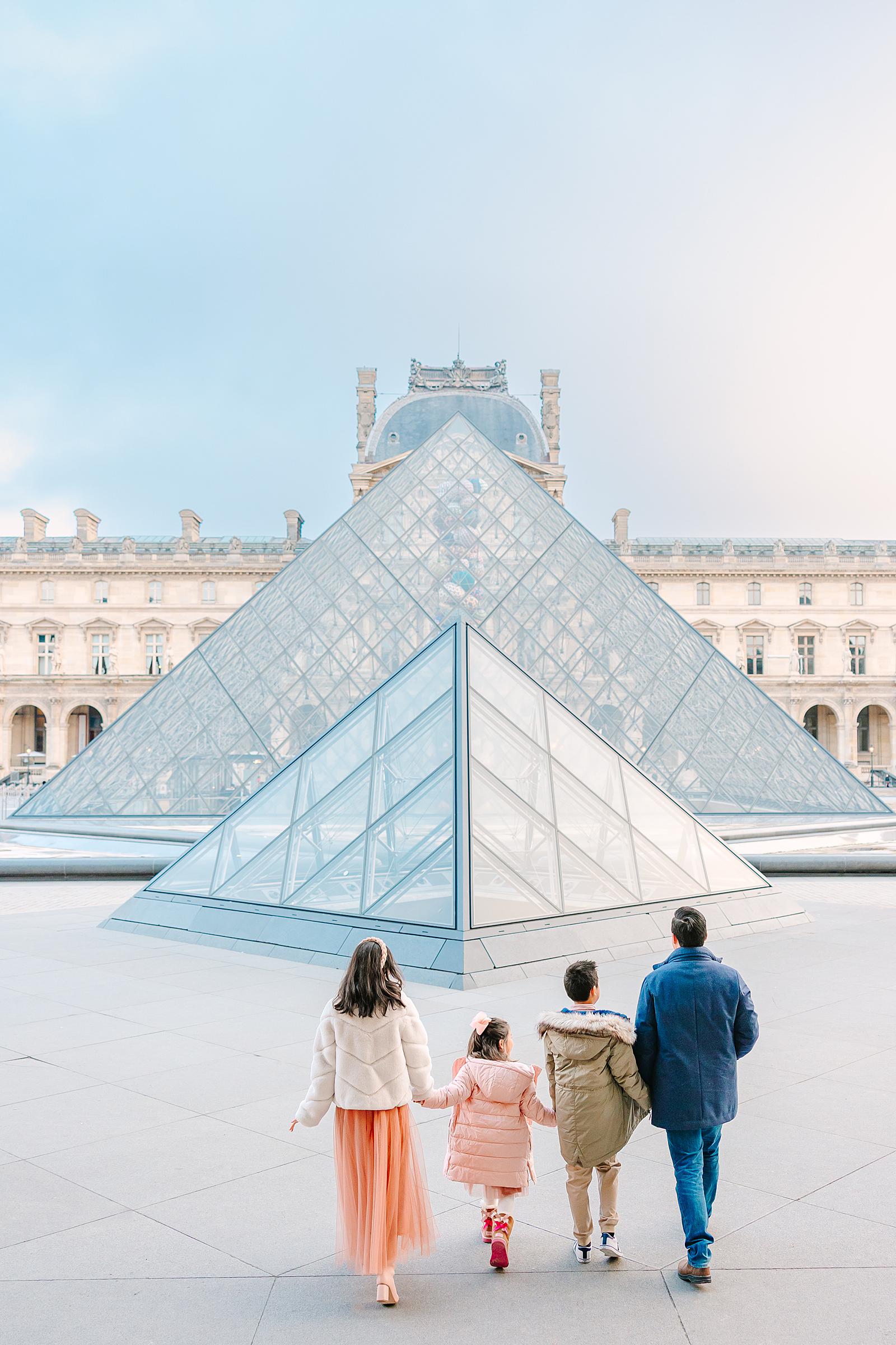 A family of four, seen from behind, walking towards the iconic glass pyramid of the Louvre Museum under a soft sky.