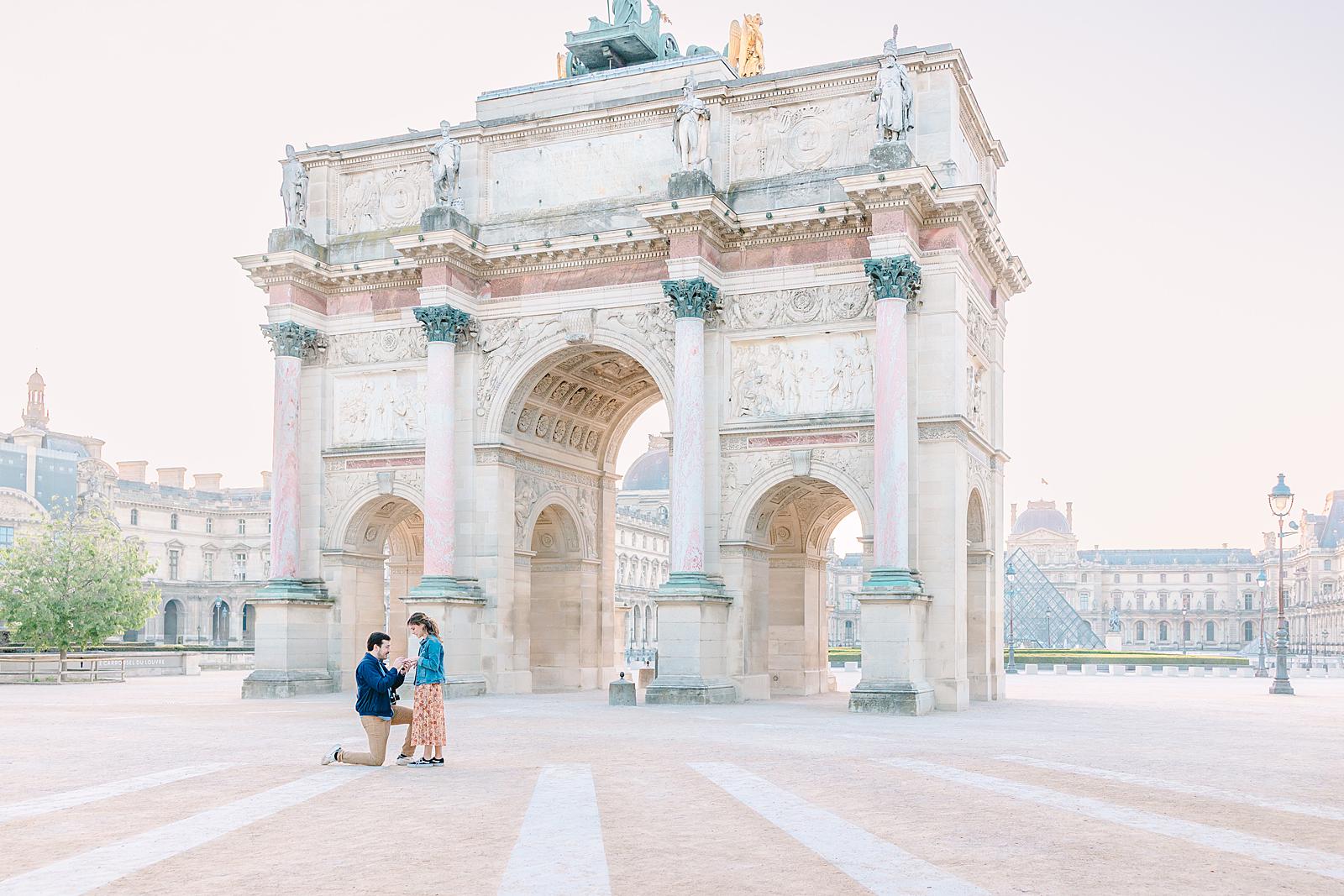 A man proposing on one knee to a woman in the Tuileries Garden, with the Arc de Triomphe du Carrousel and the Louvre Pyramid in the background.