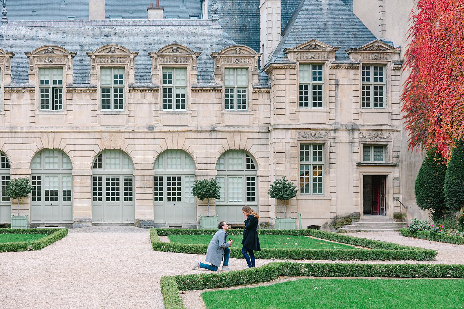 A man is down on one knee proposing to a woman in the beautifully manicured gardens of Hôtel de Sully, with the grand palace facade in the background.