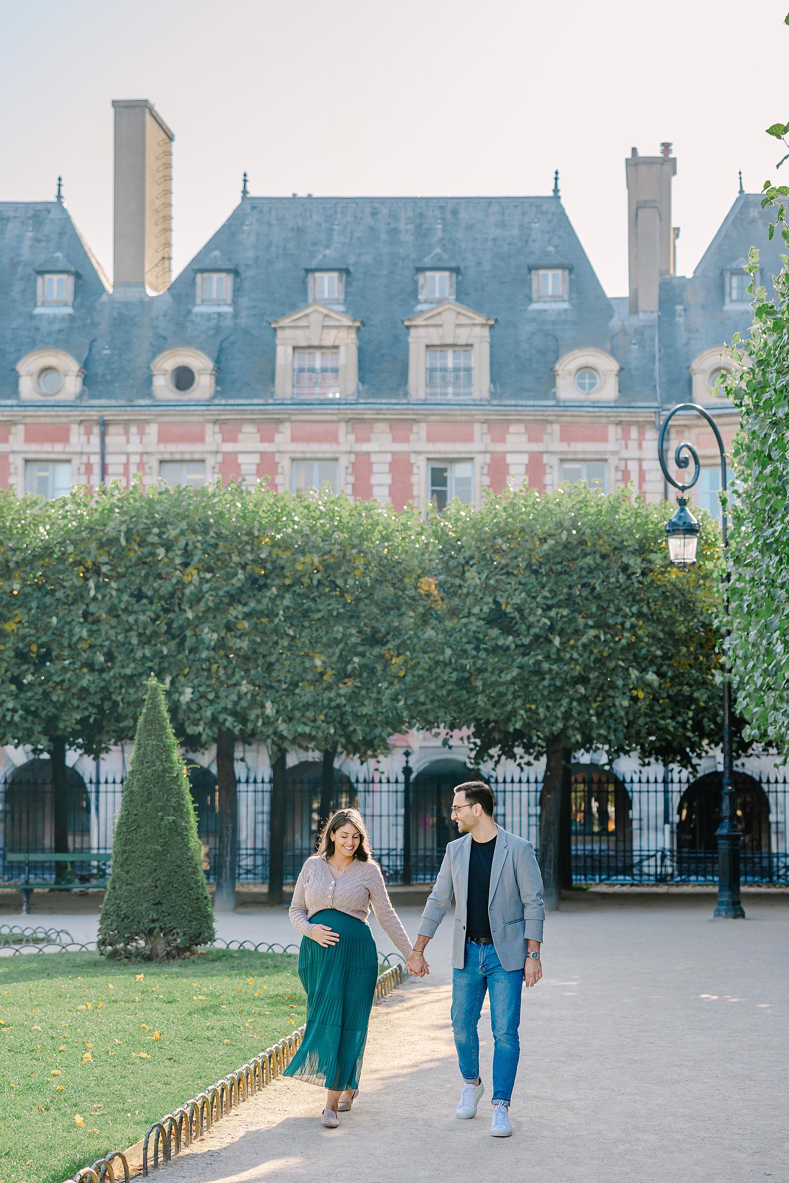 Expecting couple holding hands and walking on a path in Place des Vosges, with the classic brick and stone buildings of the square behind them.