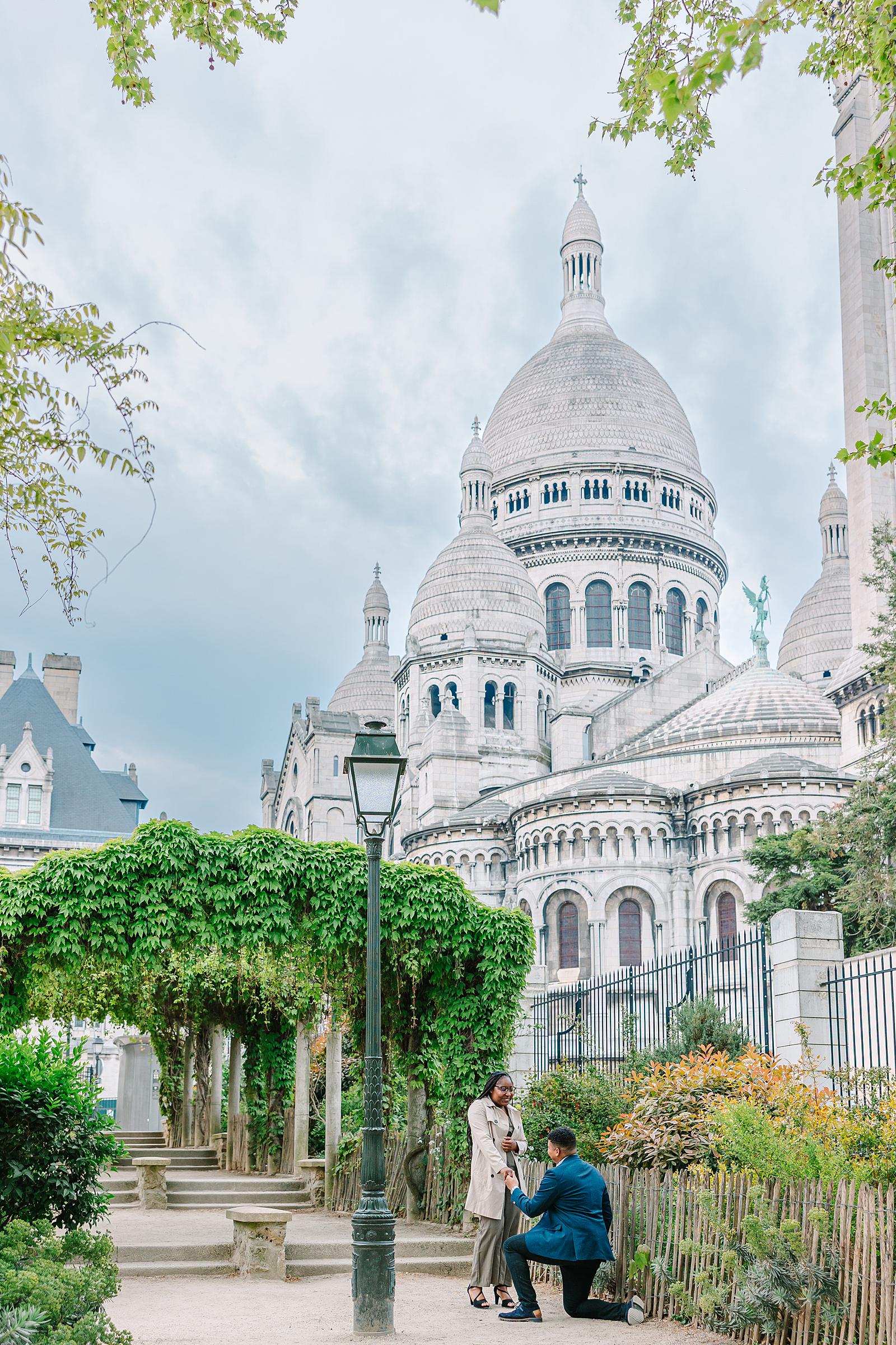 A man on one knee proposing to a woman in a lush garden setting of Parc de la Turlure, with the impressive Sacré-Cœur Basilica behind them.