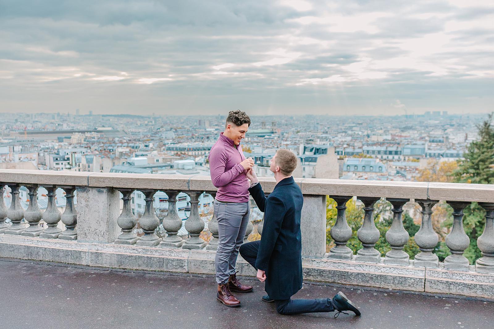 One person on one knee, holding their partner's hand, as they smile at each other during a proposal at the Montmartre viewpoint overlooking Paris.
