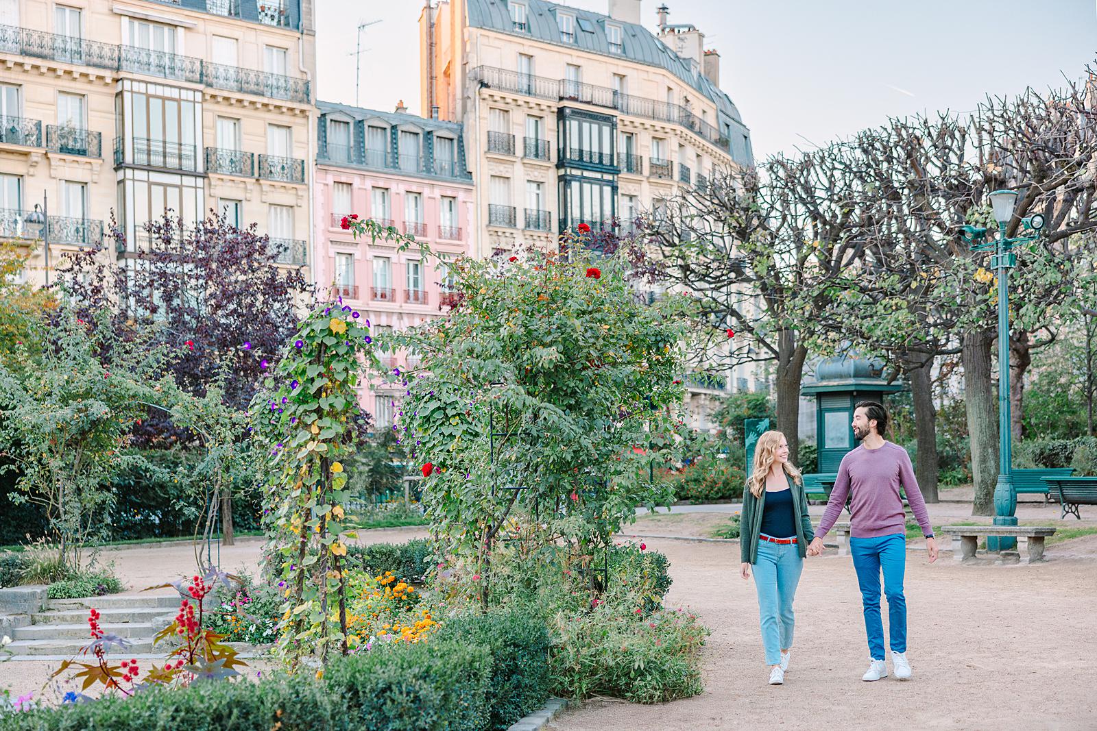 A happy couple, holding hands, walking along a path in Square René Viviani with colorful flower beds and classic Parisian buildings in the background.