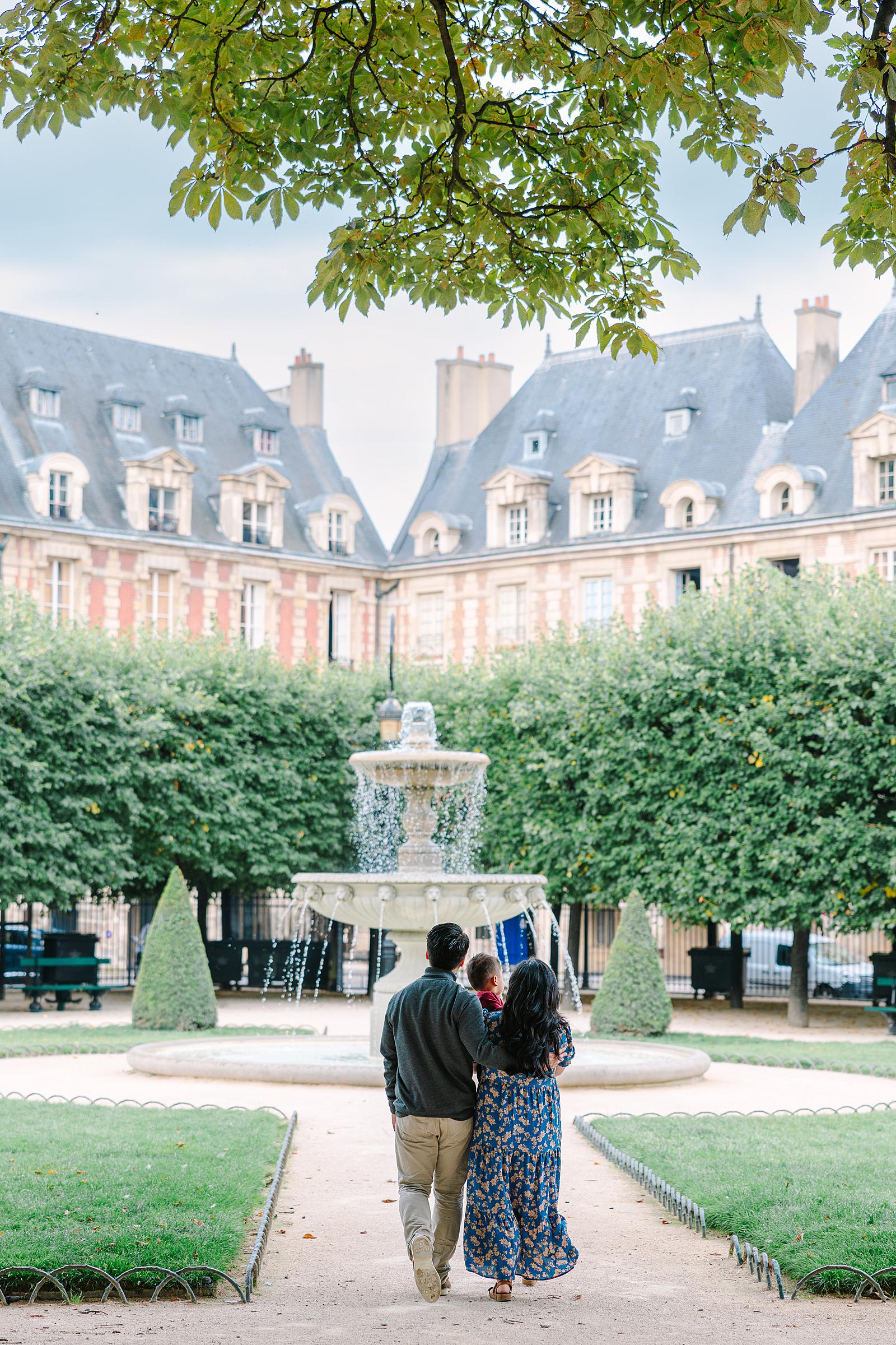 A family of three, parents holding their toddler, walking away from the camera towards a fountain in the center of Place des Vosges park, with symmetrical buildings in the background.