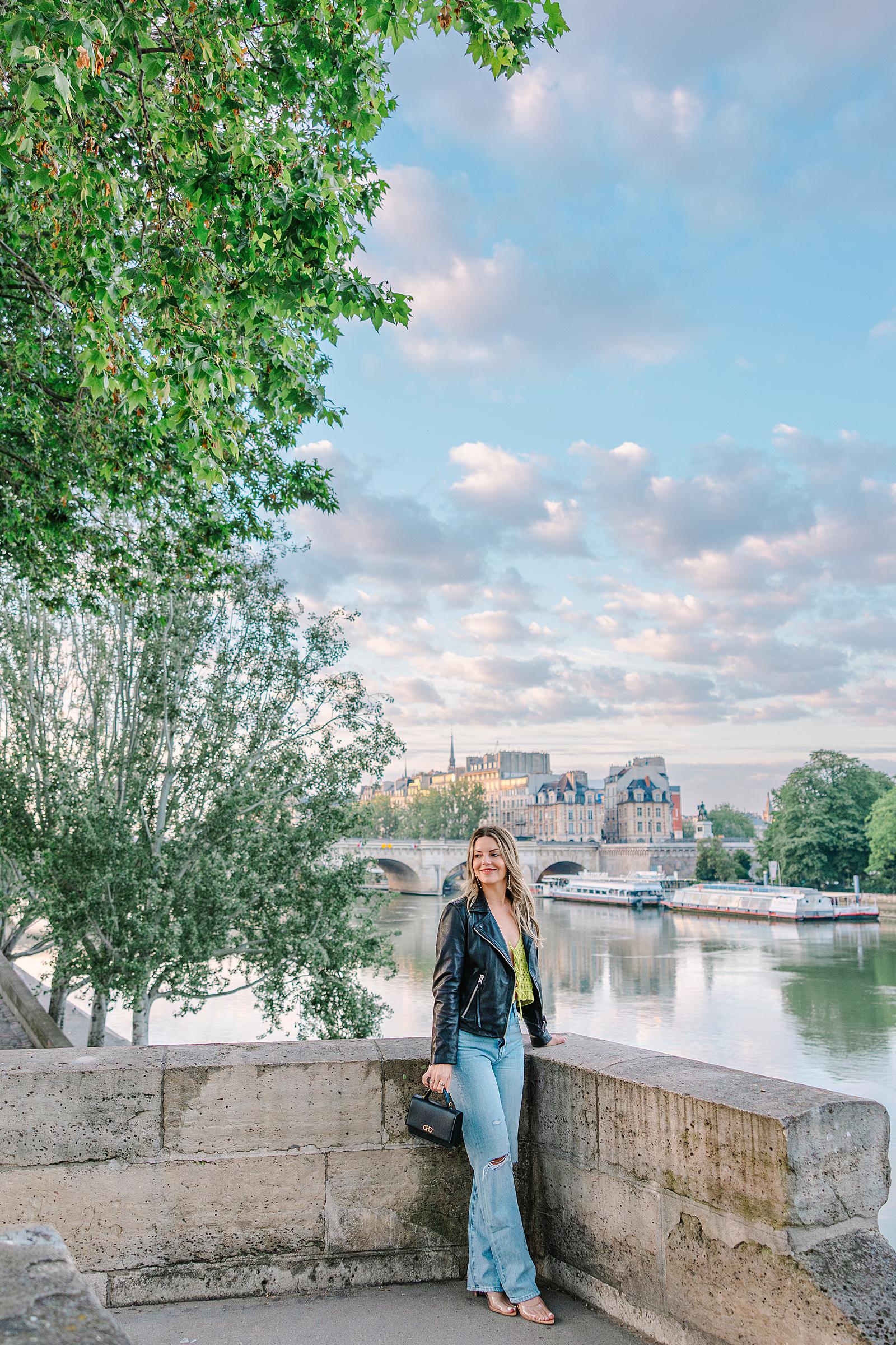 A woman in a black leather jacket and jeans standing by a stone wall near the Seine River, under green trees, with Parisian buildings and bridges in the distance.